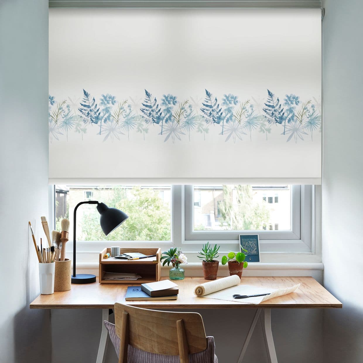 Roller blind with pale fabric and blue botanical border, lowered across the window, gently filtering daylight; small wooden desk beneath with lamp, potted plants, and stationery in a bright home workspace.