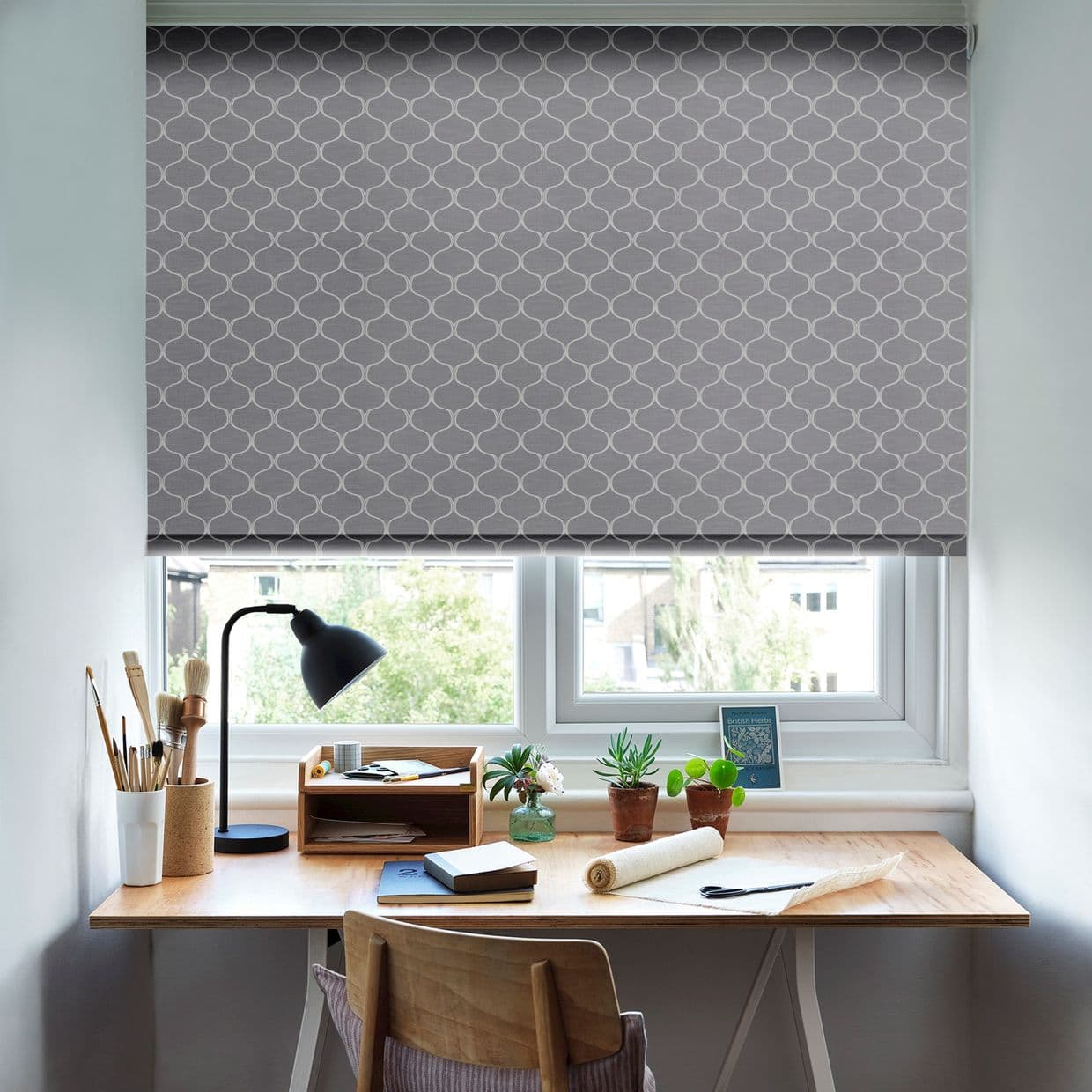 A grey roller blind with white trellis pattern lowered over a double window; it filters daylight. Below, a tidy wooden desk holds a lamp, brushes, potted plants and notebooks.