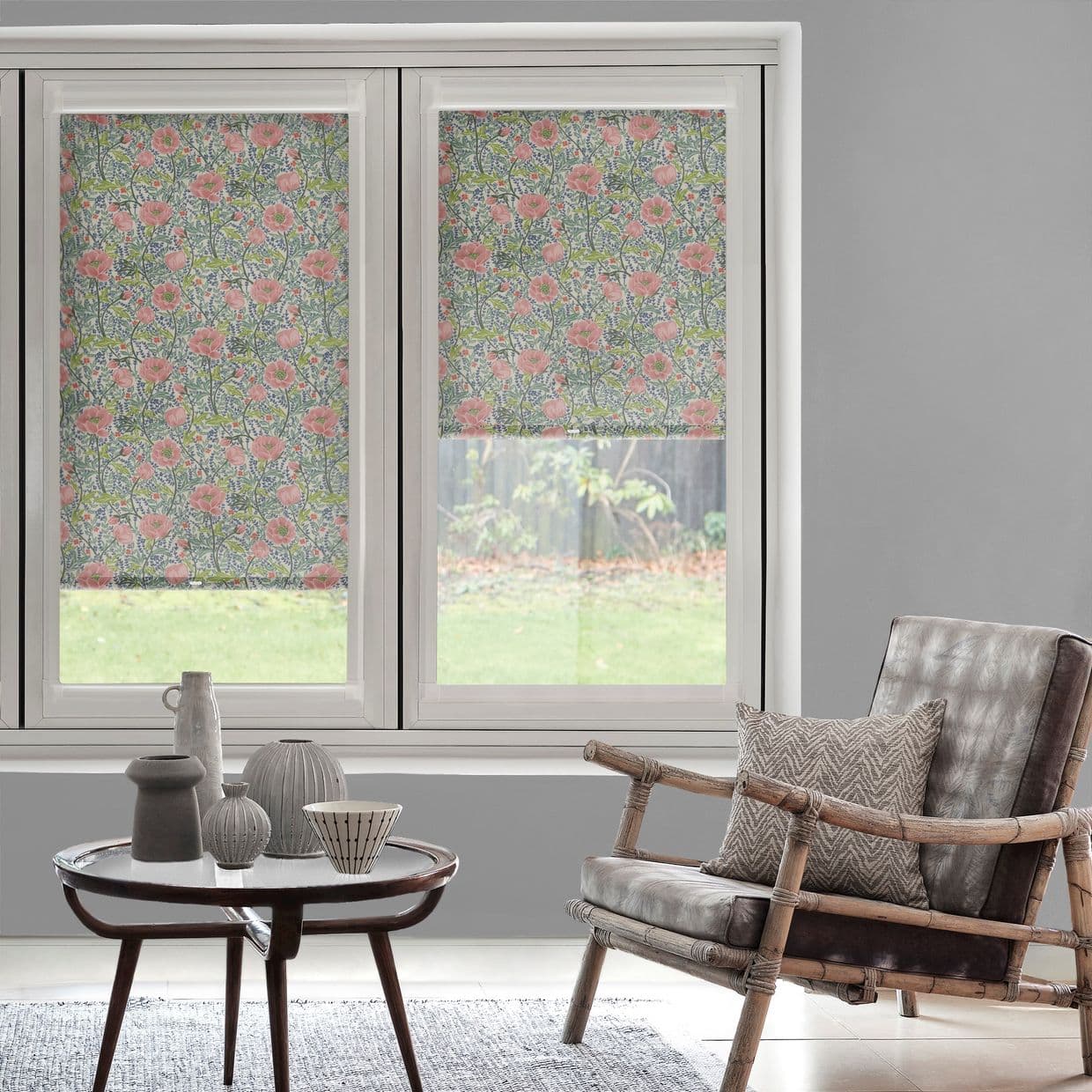 Floral patterned roller blinds (pink poppy motif) on dual windows â€” left fully lowered, right half-raised â€” filtering daylight into a grey-walled sitting area with rattan chair and table.