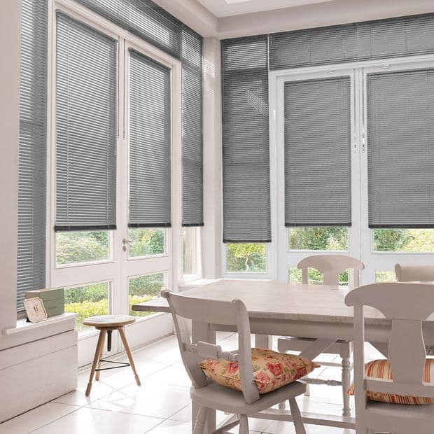 Grey horizontal Venetian blinds cover tall bay windows, fully lowered and filtering daylight; wooden dining table with chairs and floral cushions in a bright sunlit breakfast nook overlooking green garden.