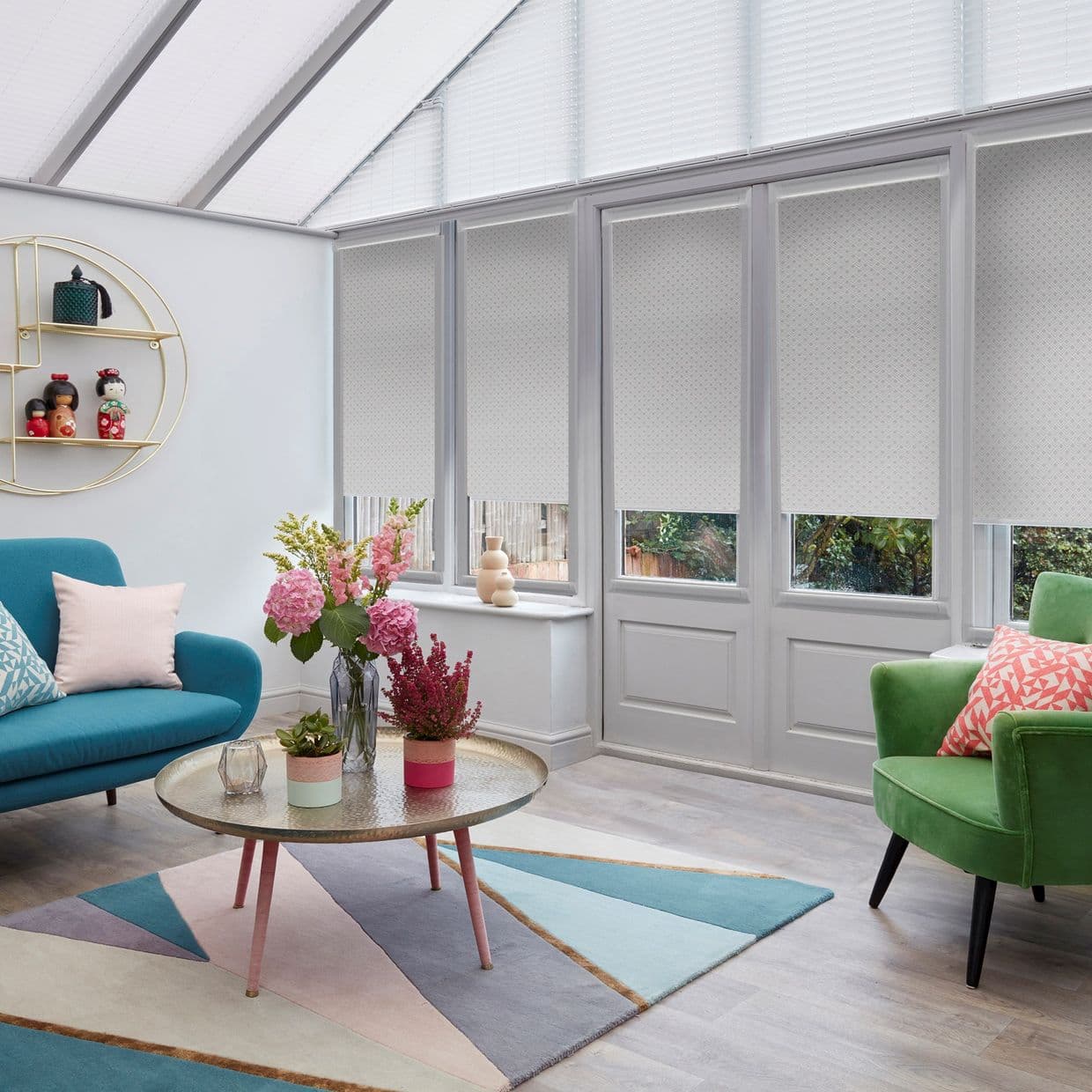 White roller blinds covering tall conservatory windows, lowered to mid-height and diffusing daylight; modern sitting room with colorful sofas, geometric rug, coffee table, and a leafy garden view beyond.