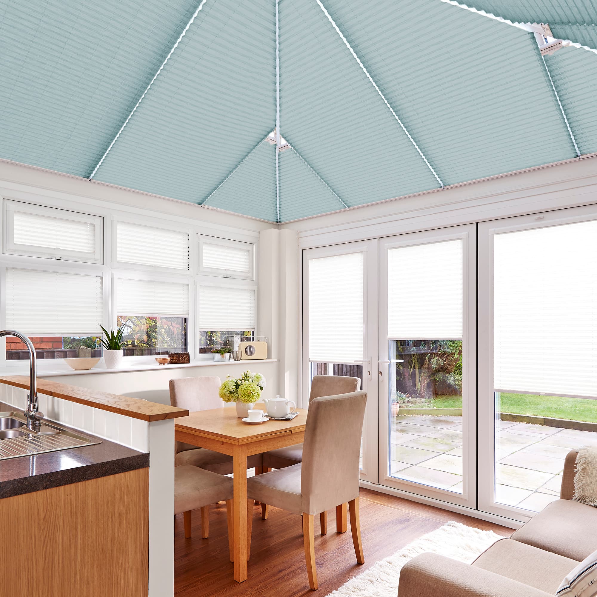 Object: pale teal pleated cellular blinds covering the pitched conservatory roof and white pleated blinds on windows and doors. Action: closed, filtering daylight. Context: bright dining conservatory with table, chairs and garden view.