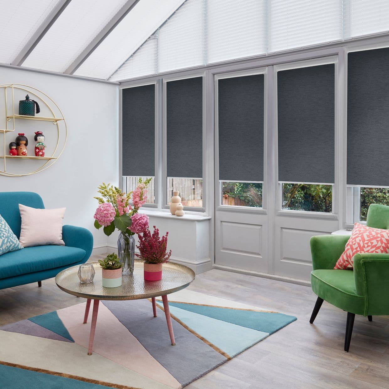 Dark grey roller blinds cover tall conservatory windows/doors, lowered over upper panes and blocking light while lower panels remain visible; bright sunroom with teal sofa, green armchair, rug, coffee table.
