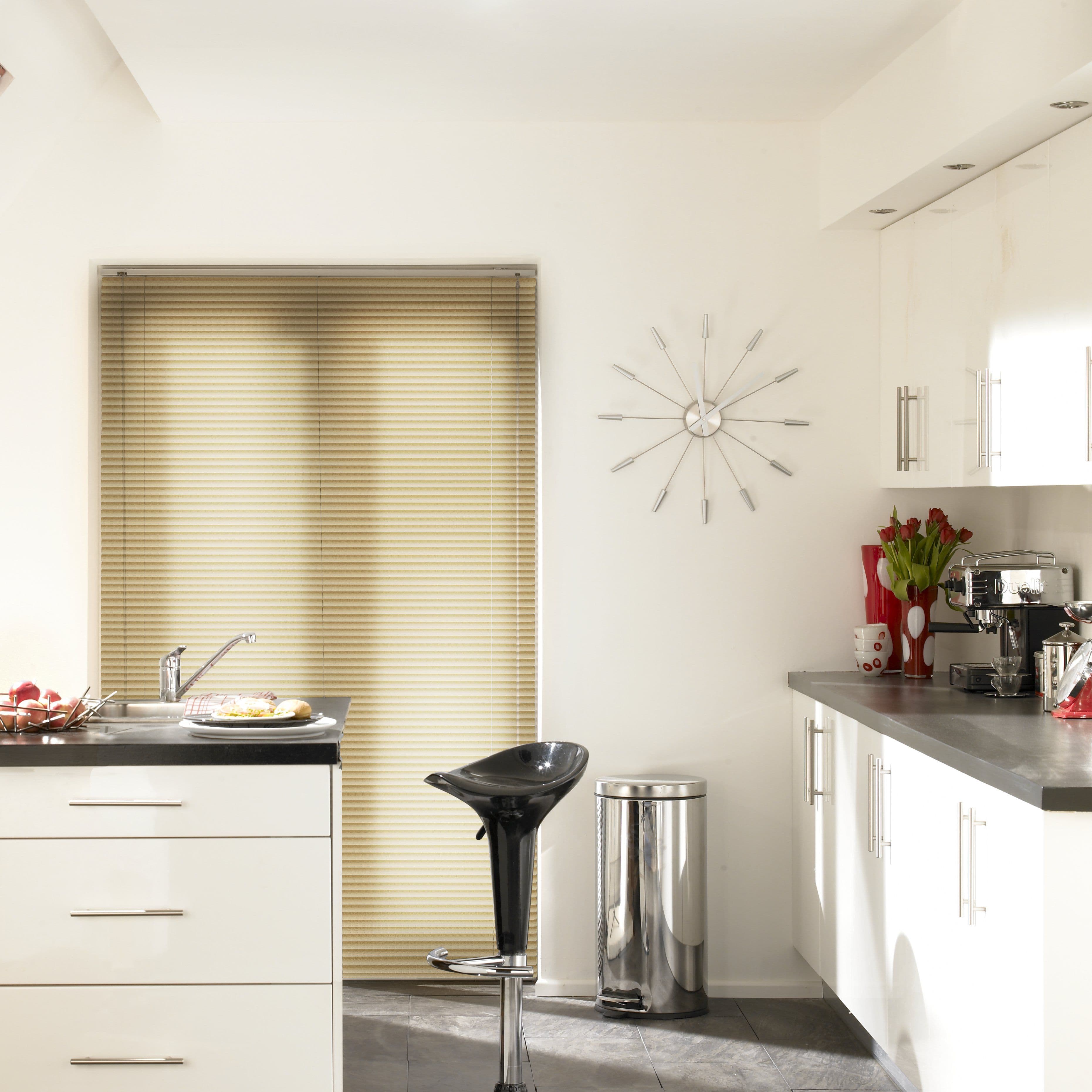 Beige horizontal Venetian blinds covering a full-height window, closed and softly filtering daylight; modern white kitchen with black countertop, glossy barstool, stainless pedal bin, wall clock, and coffee machine.