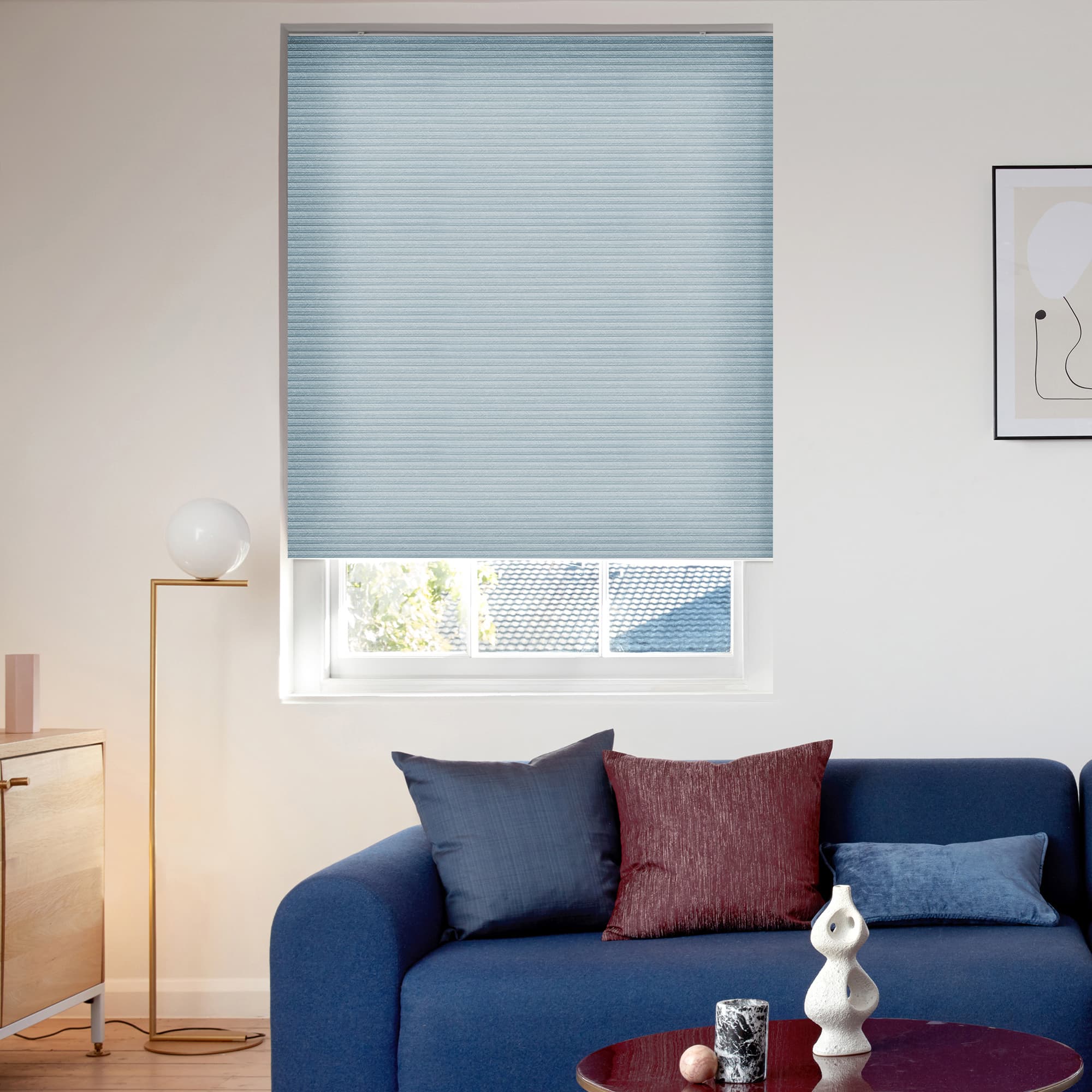 Light-blue pleated blind, lowered inside the window recess with ribs; softly filtering daylight while leaving a lower gap; above a blue sofa with cushions, brass floor lamp and minimalist wall art.