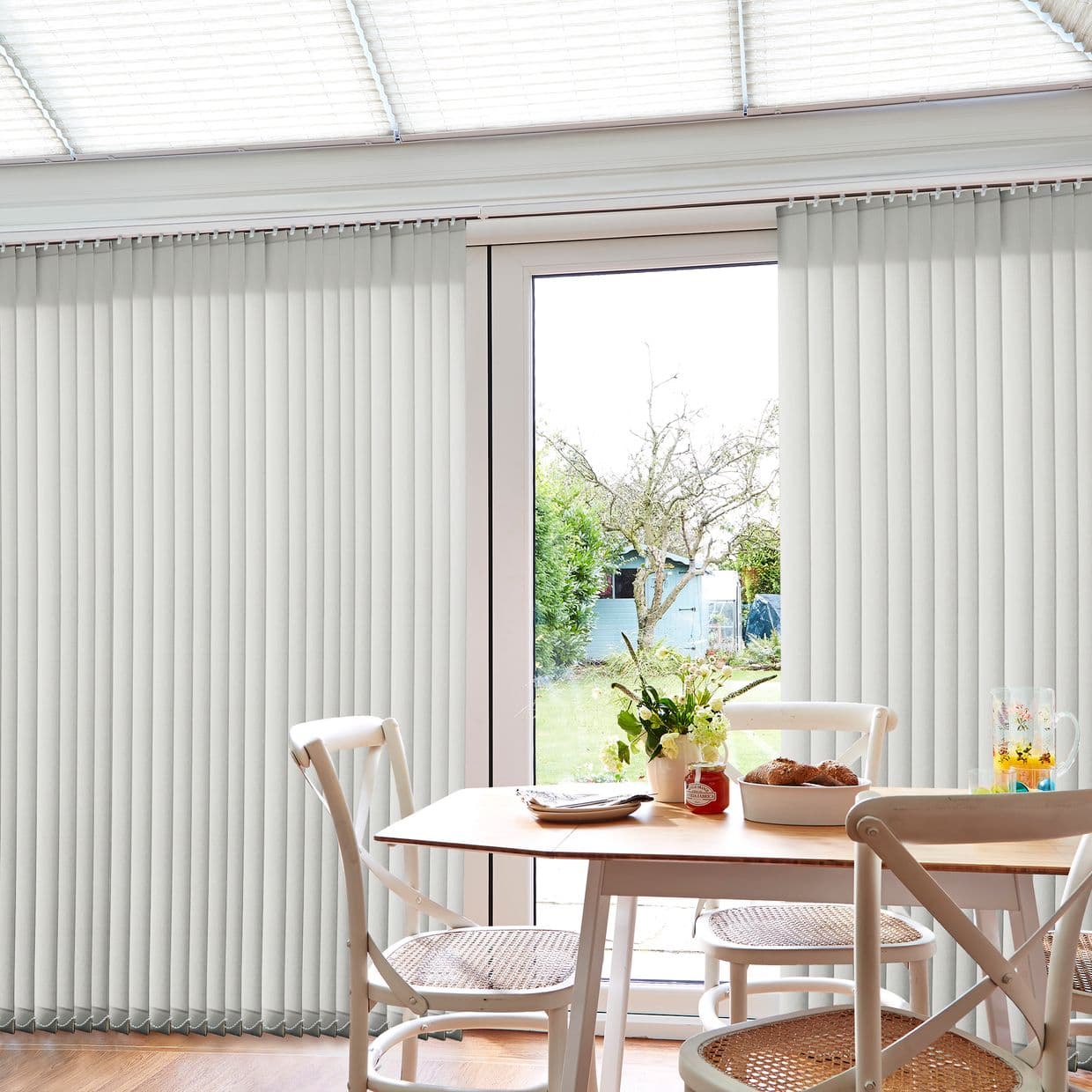 Cream vertical blinds, floor-to-ceiling panels split to reveal a central glazed patio door, partially open; filtering daylight into a dining area with wooden table, wicker chairs and a garden view.