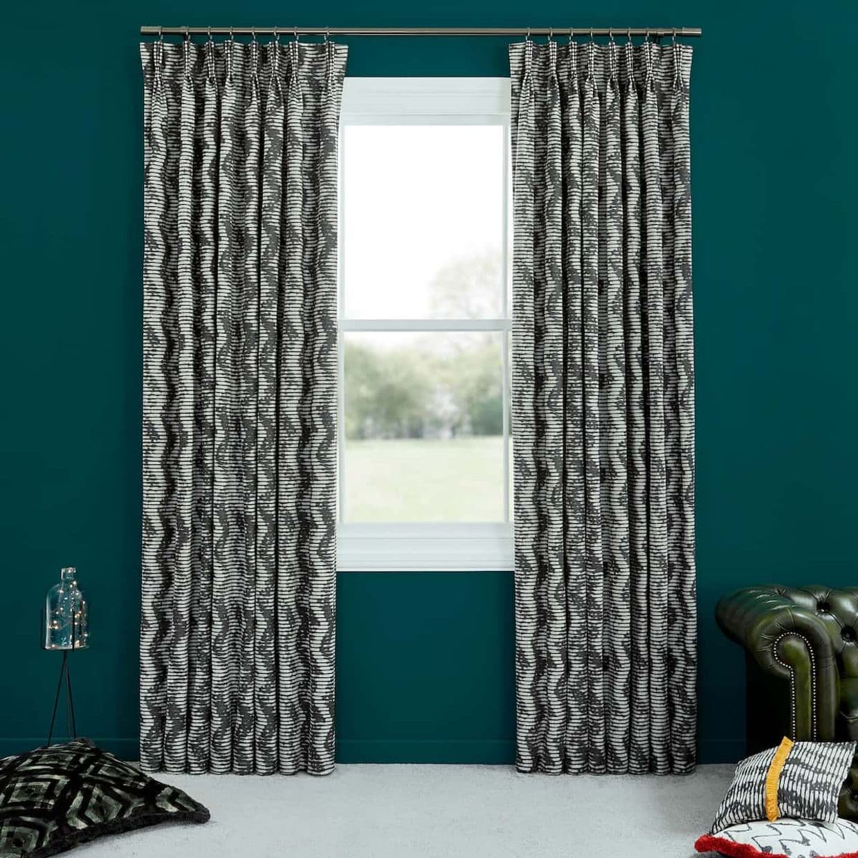Black-and-white wavy-striped pleated curtain panels, hung on a metal rod and parted, framing the central window and admitting daylight into a teal-walled room with cushions and a green leather armchair.