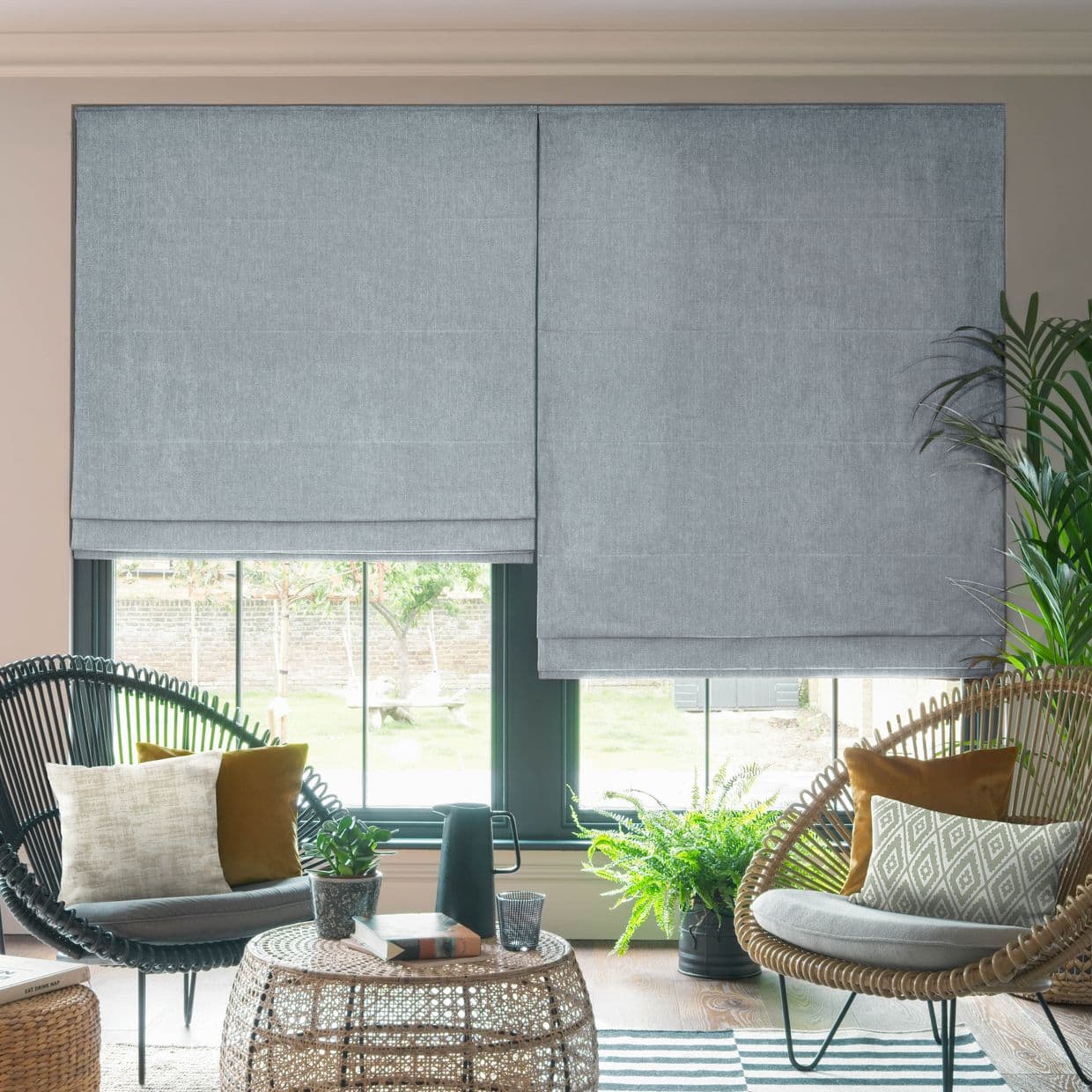 Light-grey textured Roman blinds, two adjacent panels with seams, cover the windows; one panel slightly raised, both filtering daylight; cozy sunlit seating with rattan chairs, plants, wicker table, garden view.