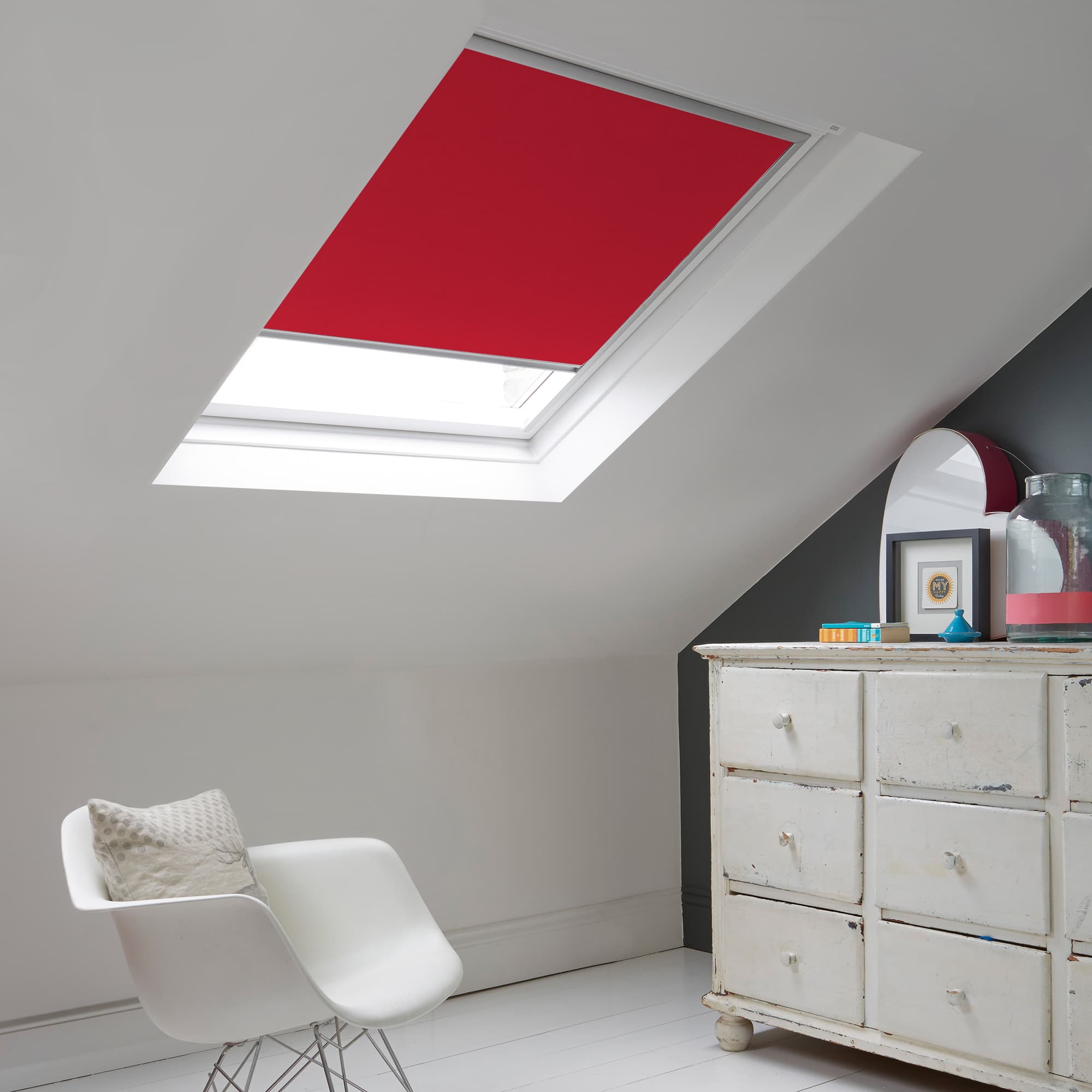 Room with white eaves wall, dark blue-grey wall to right with a shabby chic white drawer unit, a contemporary white chair, white painted floorboards and a skylight blind in pale red.