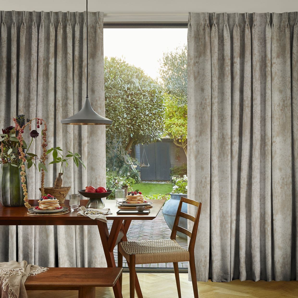 Floor-length pleated grey-beige curtains with deep folds, parted centrally to reveal a glass door; filtering soft morning light over a wooden dining table set with pancakes and a garden.