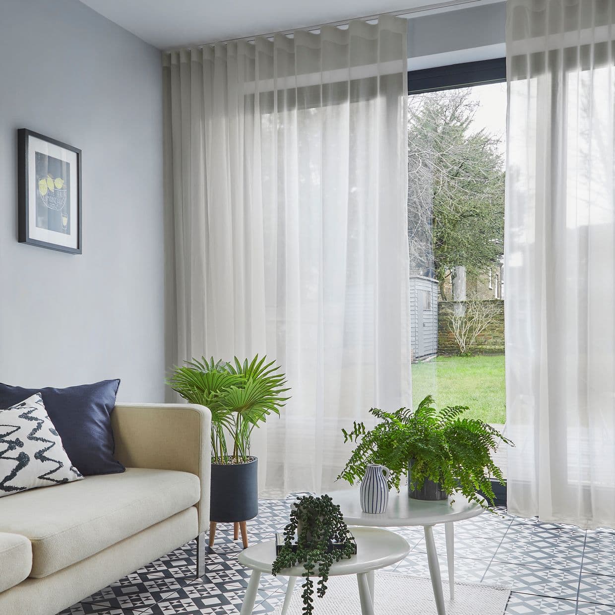 Sheer floor-to-ceiling white curtains hang across a sliding glass door, softly filtering daylight; in a bright living room with beige sofa, potted plants and patterned tile floor overlooking a lawn.