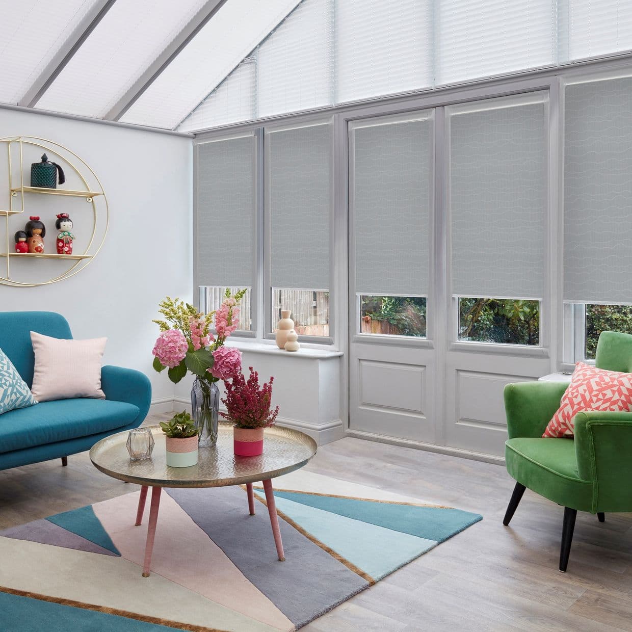 Grey patterned roller blinds cover tall windows, lowered to reveal narrow clear bottom panels and softly filter daylight; bright living room with teal sofa, green armchair, coffee table with flowers.