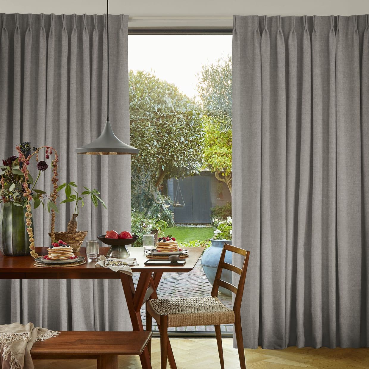 Gray floor-to-ceiling pleated curtains, slightly parted in the center with soft folds, revealing a sunlit backyard; dining table with breakfast settings, pendant lamp and wooden chair in foreground.
