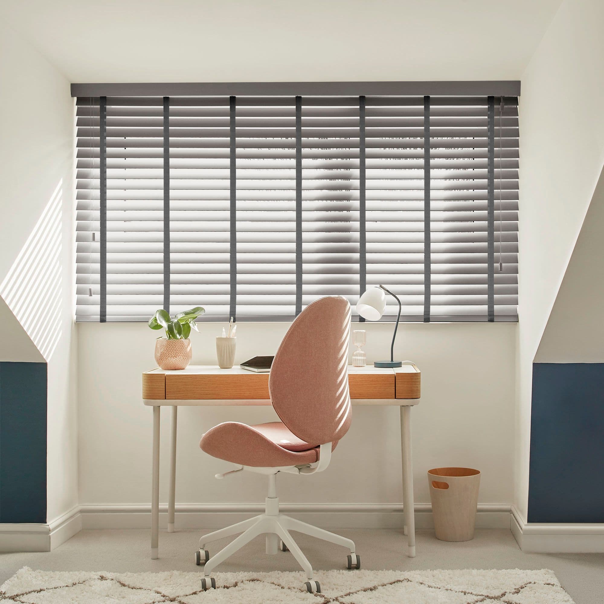 Grey horizontal slatted blinds covering a wide dormer window, slightly tilted to filter sunlight; above a desk in a bright attic study with a pink swivel chair, lamp and plant.