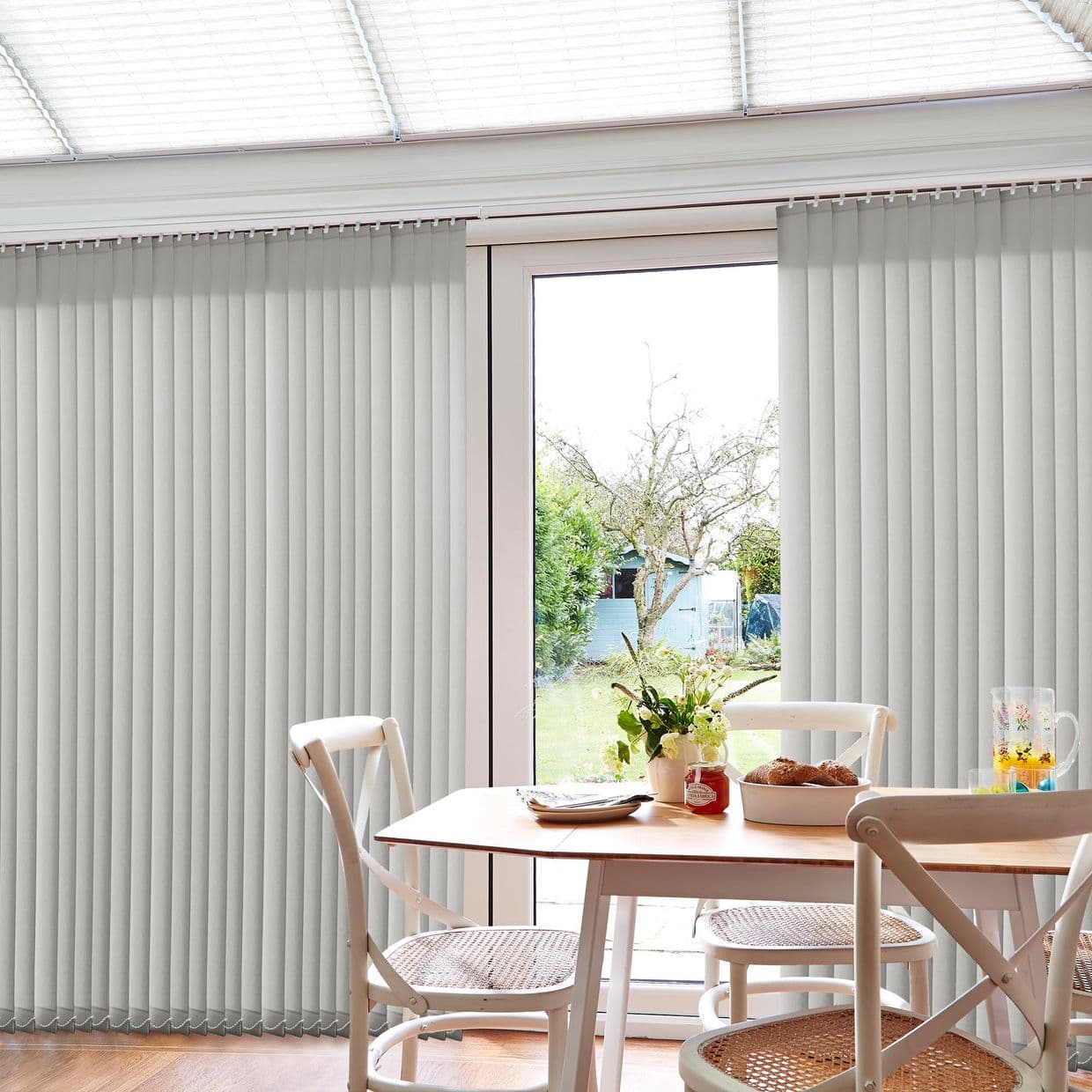 Vertical fabric blinds over patio doors, slatted and mostly closed with center parted, filtering warm daylight and revealing a garden; wooden dining table, chairs, vase of flowers and pastries.