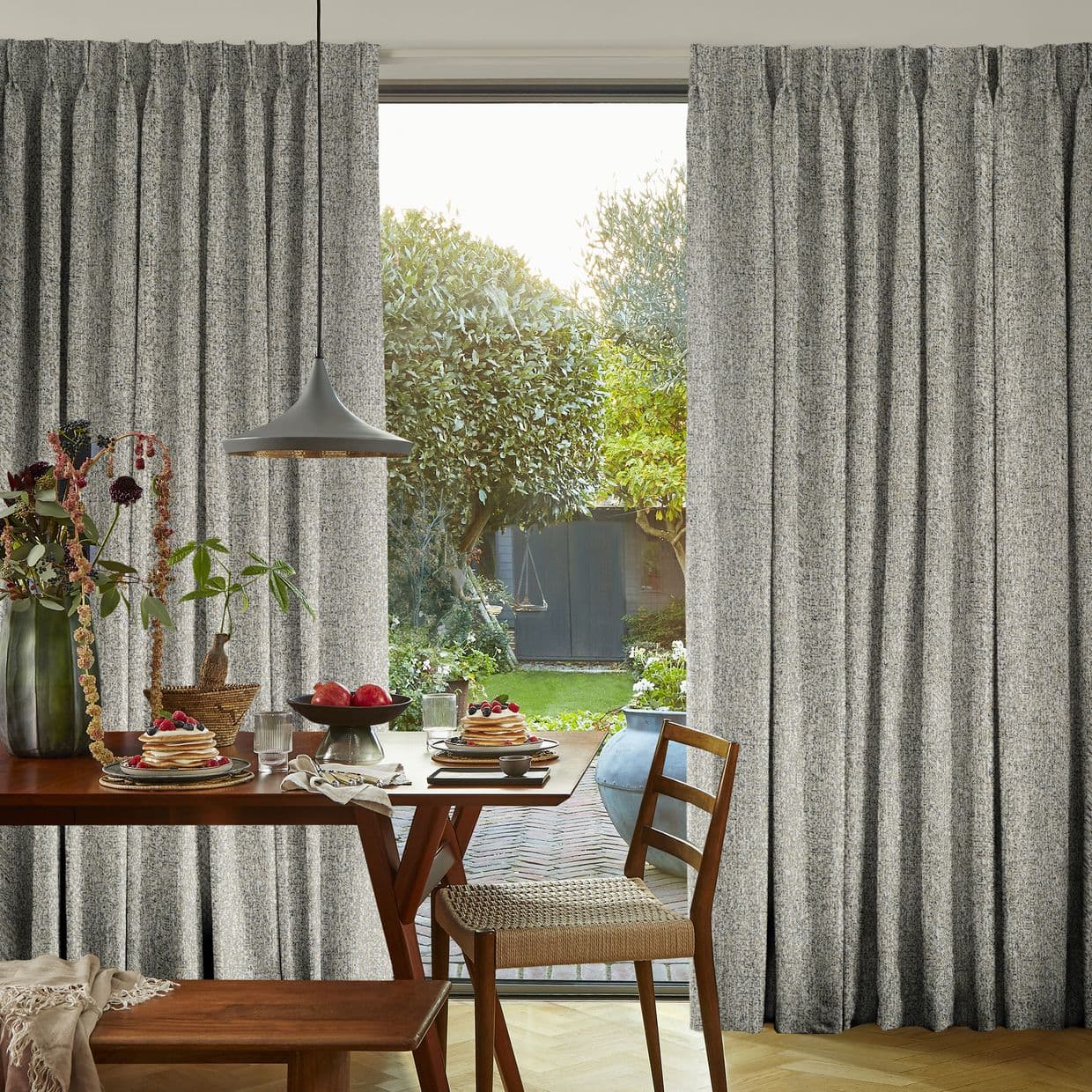 Grey textured pleated floor-length curtains, parted at the center to frame and filter daylight; a wooden dining table with plates and a pendant lamp sits before a sunlit garden.