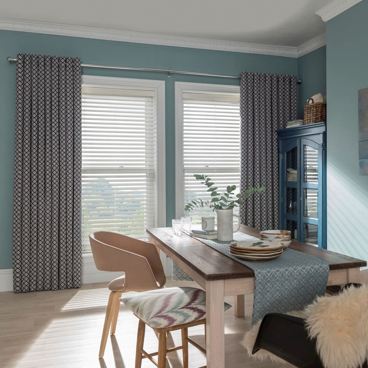 Patterned drapes on a rod flank two tall windows; lowered white horizontal blinds filter daylight in a sunlit teal dining room with wooden table, chairs, table runner and a blue cabinet.