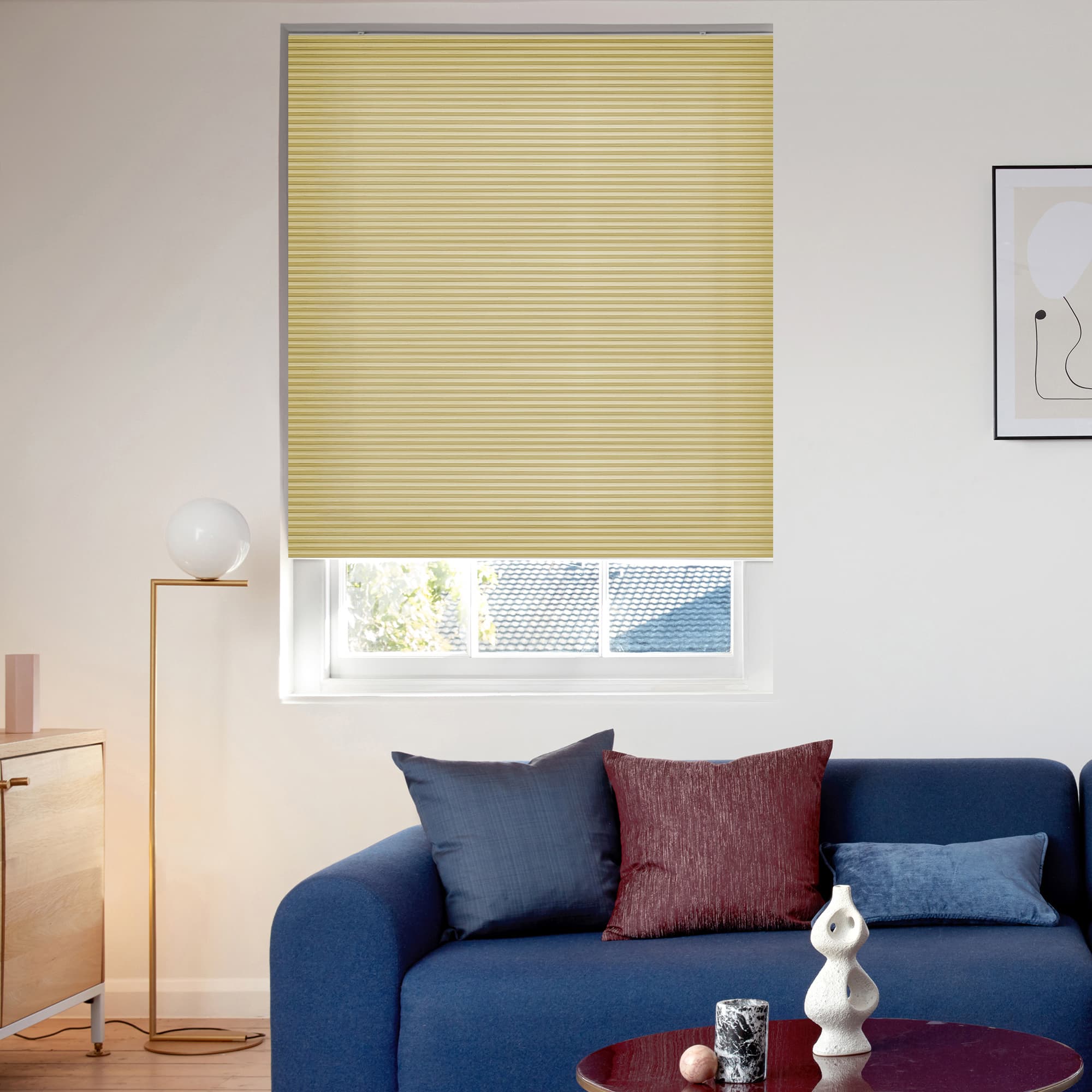 A pale-yellow pleated cellular blind, lowered to cover most of the tall window with a narrow lower gap, filtering daylight in a bright living room above a blue sofa.