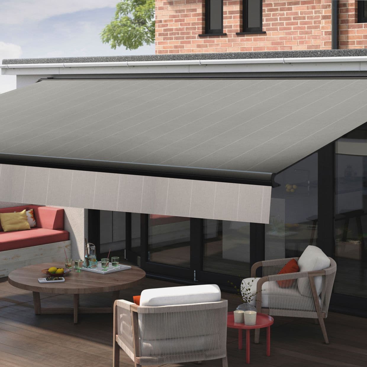 A grey retractable fabric awning with a light valance, extended over glass patio doors, shading a modern wooden terrace with lounge chairs, coffee table and brick-backed house in daylight.