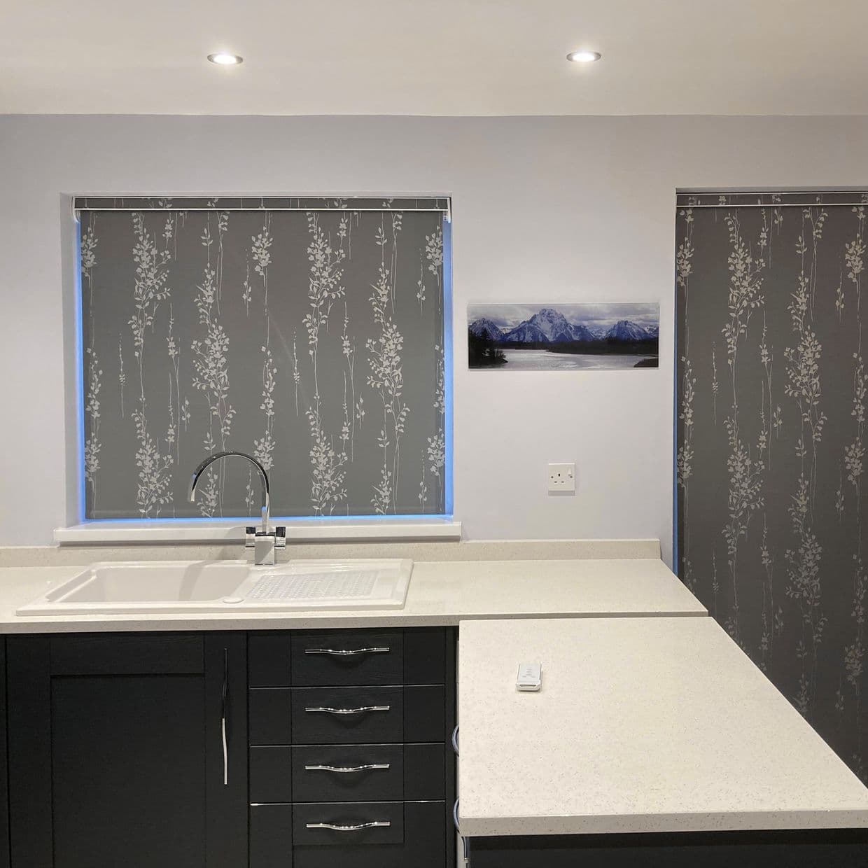 Grey roller blinds with white vertical floral pattern, fully lowered blocking light; over kitchen windows above a white sink and countertop, dark cabinets, recessed ceiling lights, small mountain print.