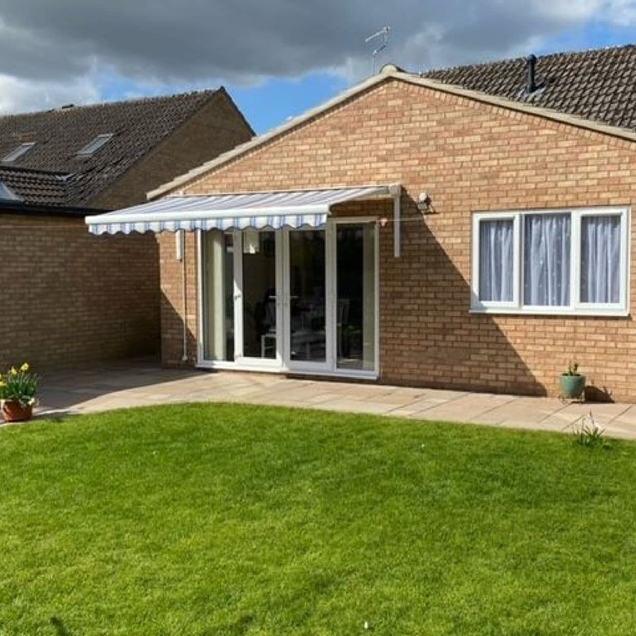 Striped blue-and-white retractable awning over patio doors, fully extended with scalloped edge, shading the glass doors. Context: single-story brick bungalow rear, paved patio and green lawn under partly cloudy daylight.