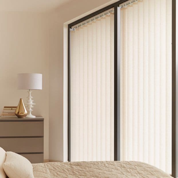 Vertical fabric vertical blinds covering two tall sliding glass doors, closed and softly diffusing daylight; beige bedroom with quilted bed, bedside dresser, lamp and decorative gold vase.