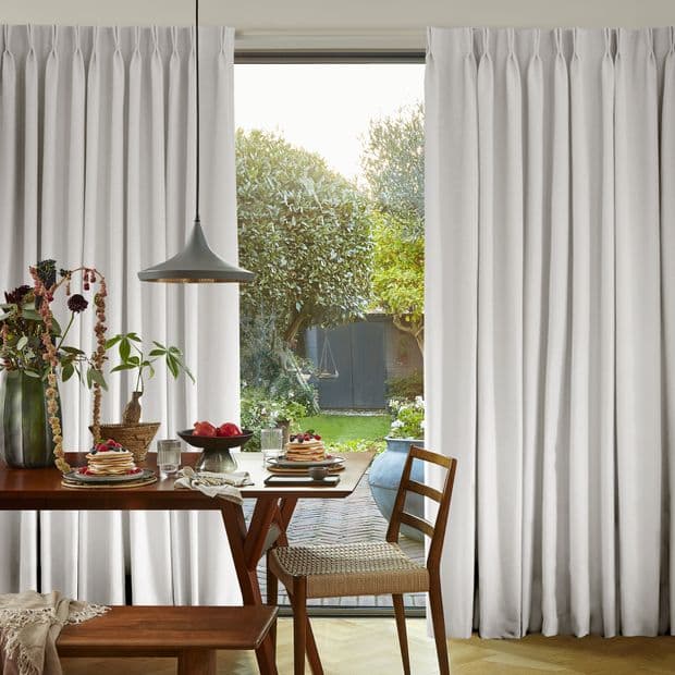 Floor-length pinch-pleat curtains, drawn apart at center to frame glass door, filtering daylight into a dining room with wooden table, pendant lamp, breakfast plates and leafy backyard view.
