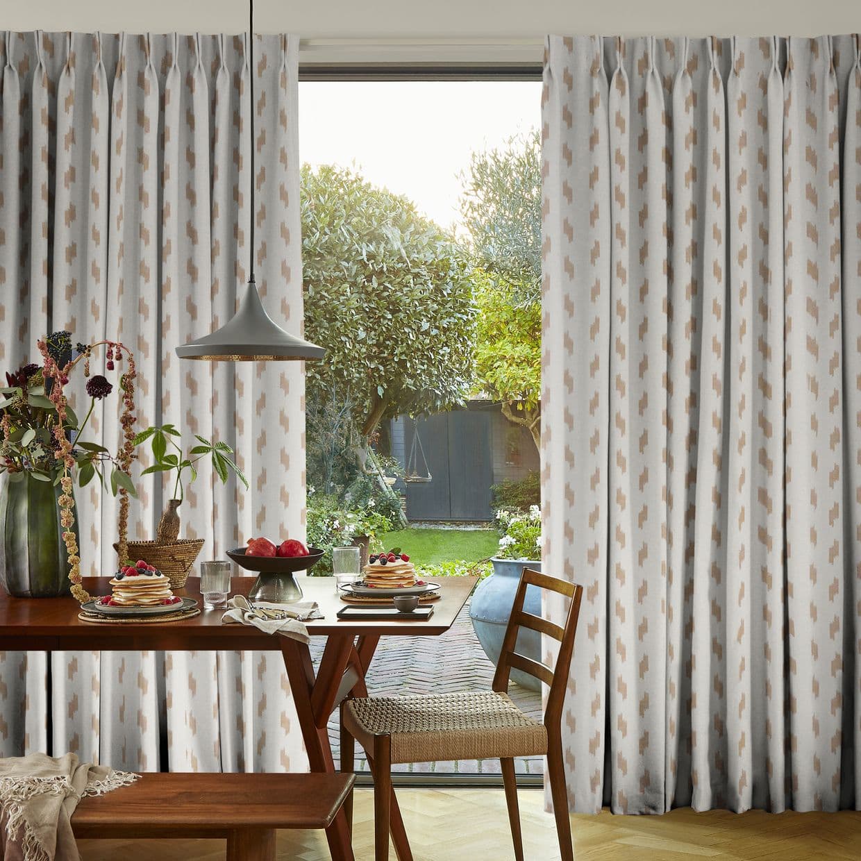 Pleated floor-length patterned curtains (cream with tan geometric motifs) parted at center, framing sliding glass door; filtering morning light over a dining table set for breakfast and green backyard.