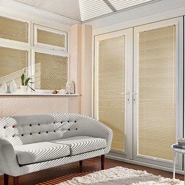 Beige horizontal blinds cover French doors and windows, fully closed and filtering sunlight, casting striped shadows across a cream tufted sofa, windowsill plants, wooden floor and white rug.
