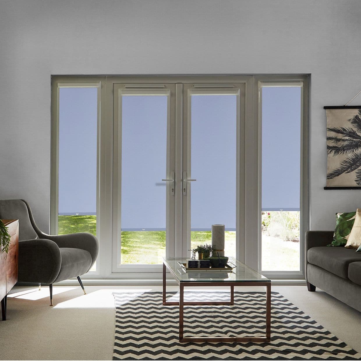 Light-blue roller blinds fitted inside the four French-door and sidelight frames, lowered to mid-height leaving a narrow bottom view and filtering light; living room with sofas, glass coffee table, rug.