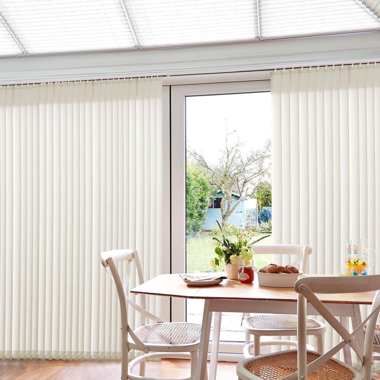 Vertical cream-coloured fabric vertical blinds covering sliding glass doors, panels partially parted at center; filtering bright daylight into a small dining area with wooden table, chairs, and a garden view.