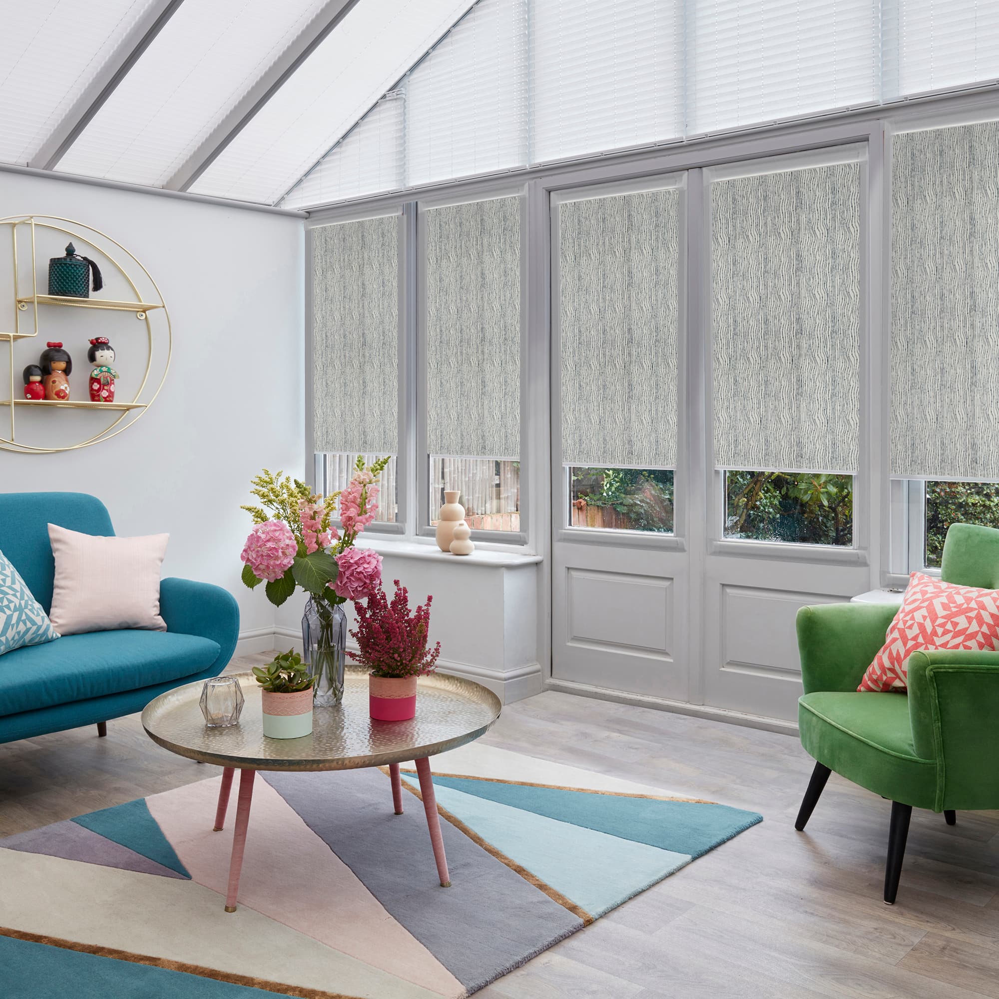 Light-grey textured fabric roller blinds lowered over the upper two-thirds of tall windows and French doors, filtering daylight into a bright conservatory-style living room with colorful sofas and coffee table.