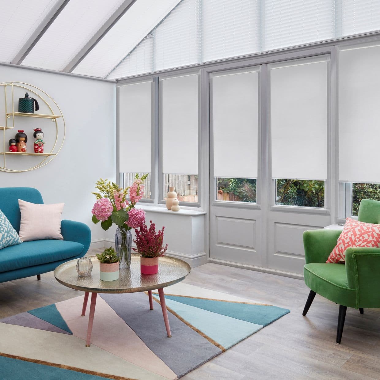 White roller blinds, lowered over tall windows, filtering soft daylight; in a bright conservatory-style living room with colorful sofas, a coffee table and floral arrangements, a garden visible outside.