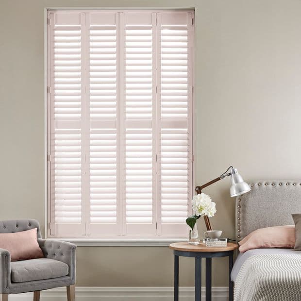 Four-panel pale-pink plantation shutters, full-height with adjustable louvers, closed and softly diffusing daylight; bedroom corner with upholstered bed and headboard, bedside table lamp and vase, tufted armchair.