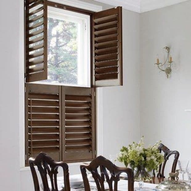 Dark wooden tier-on-tier interior shutters, upper panels open and lower panels closed, louvered slats angled, filtering daylight into a dining room with table, chairs, floral centerpiece and wall sconce.