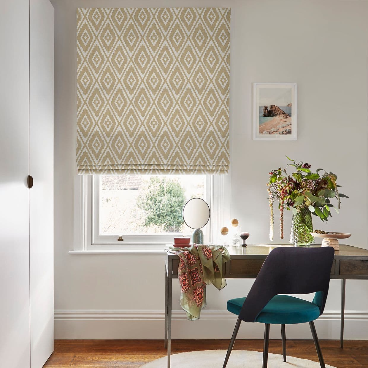 Beige-and-white diamond-patterned Roman blind lowered over a window, filtering soft daylight; a desk beneath holds a mirror, vase of flowers, scarf and teal chair in a bright, neutral home office.