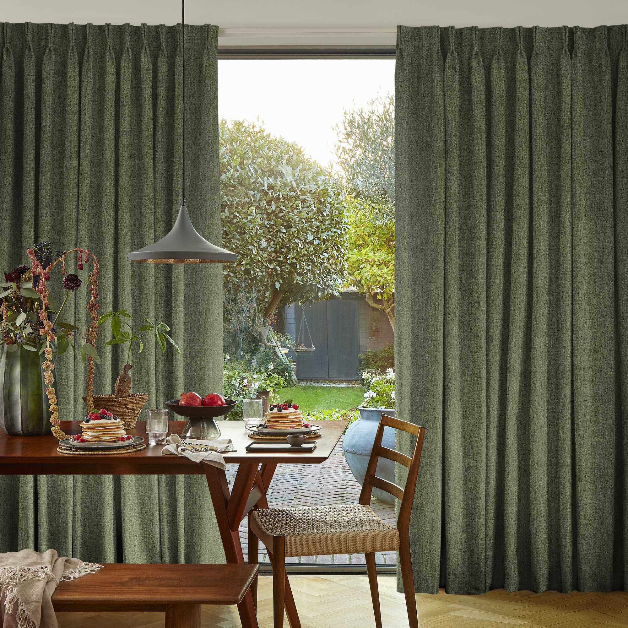 Floor-length, pleated olive-green curtains, drawn open at the center, framing a sliding glass door; they reveal a sunlit dining corner with a wooden table set for breakfast and a leafy backyard.