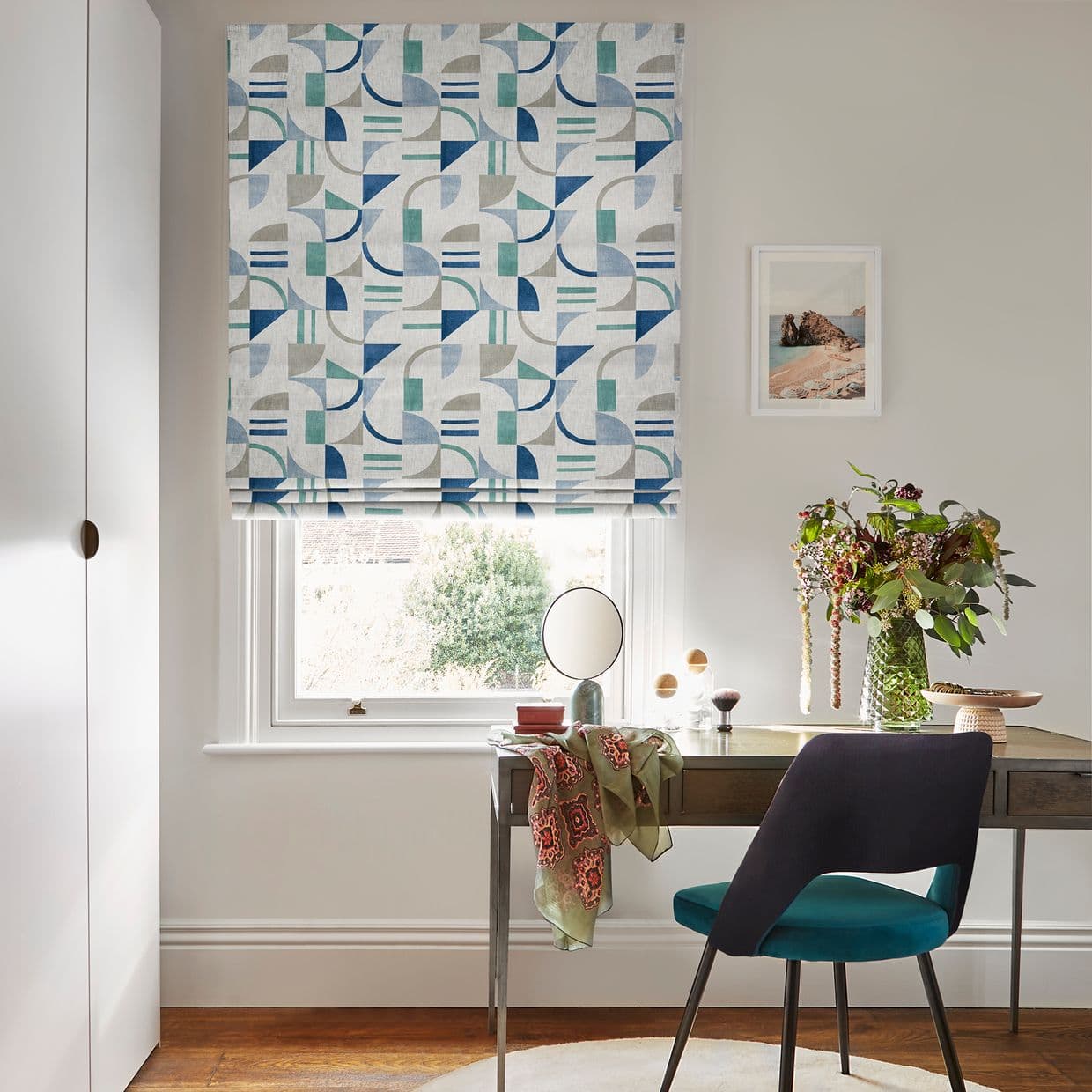 A geometric-patterned Roman blind (blue, teal, grey shapes) lowered over a sash window, filtering daylight; set above a wooden desk with chair, mirror, vase of flowers and framed coastal print.