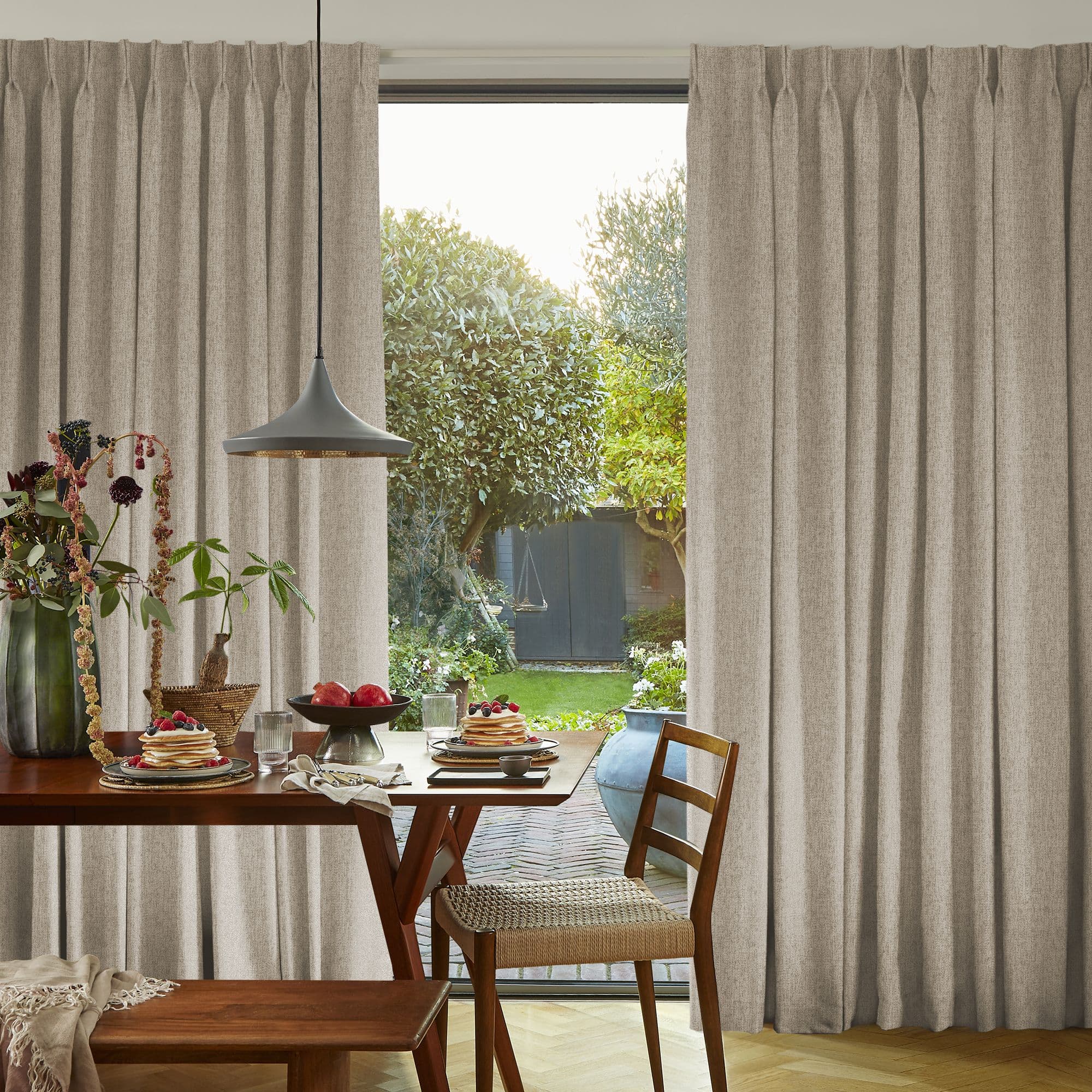 Beige floor-length pleated curtains, hung and parted at center revealing a glass door, filtering soft daylight onto a wooden dining table set for breakfast, with a green garden visible beyond.