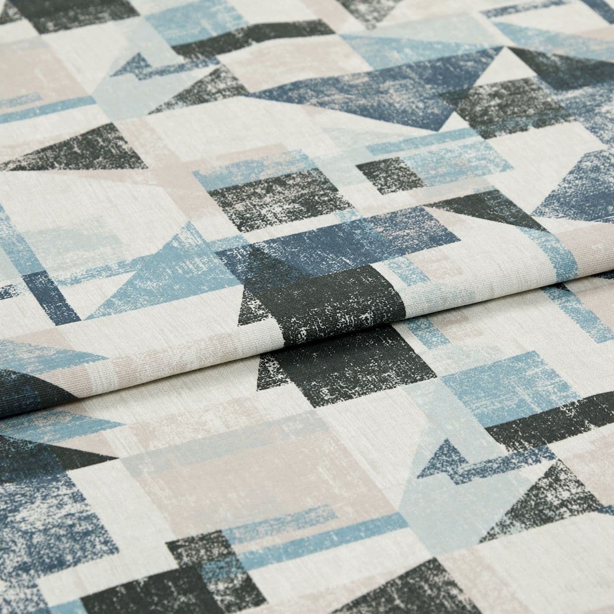 Folded geometric-print fabric sample with cream base and distressed blue, black and pale pink triangles and rectangles â€” draped flat in a close-up studio shot under soft even lighting.