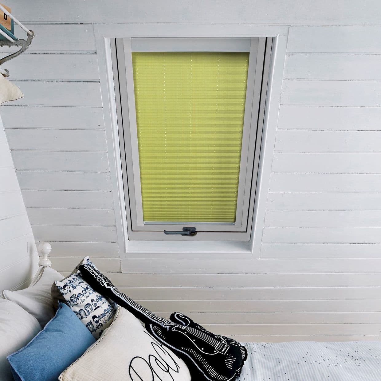 A yellow pleated blind, fully lowered inside a white-framed skylight, filters daylight in a white-painted attic bedroom; a bed beneath holds patterned pillows and a guitar-shaped cushion.