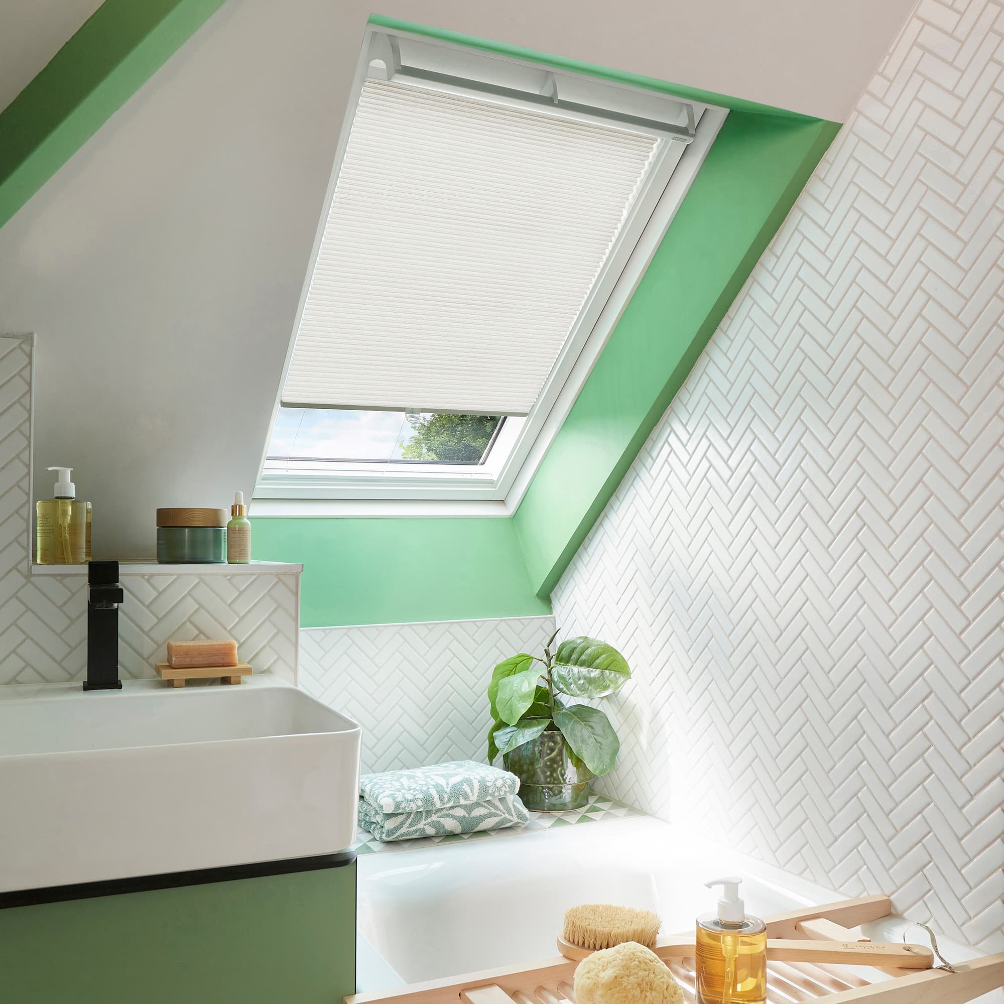 White pleated skylight blind, partially lowered across an angled roof window, filtering daylight â€” bathroom with green-painted window trim, white herringbone tiles, bathtub, sink, potted plant, and toiletries.
