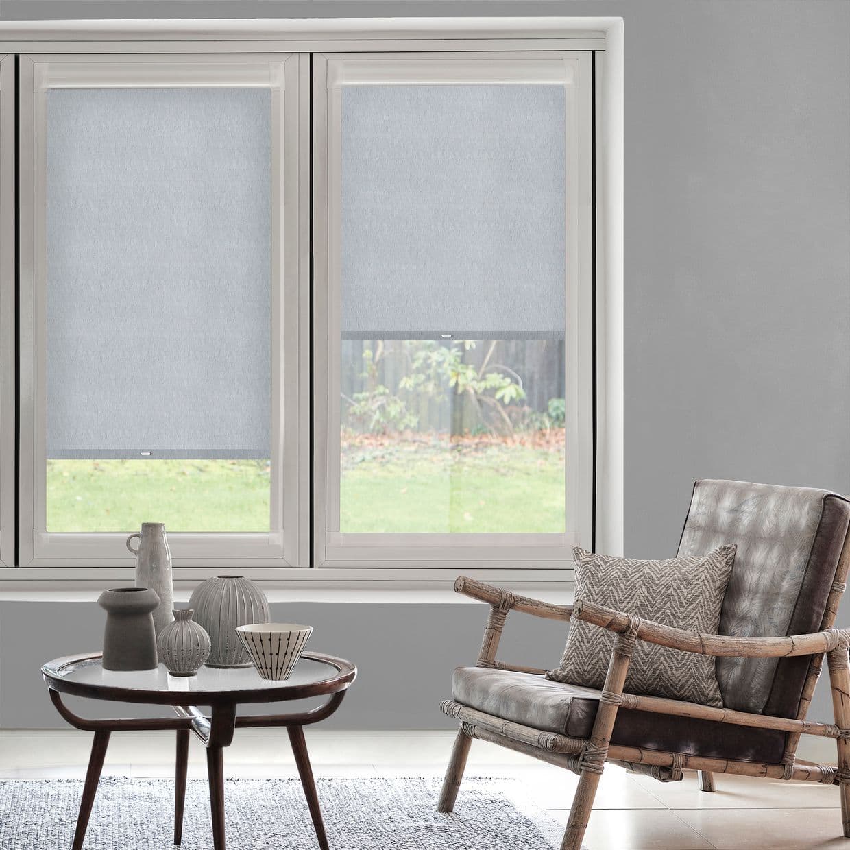 Two light-gray roller blinds covering adjacent windows, left fully lowered, right partially raised showing garden; light-filtering fabric with visible hem; modern living room with neutral tones and rattan chair.