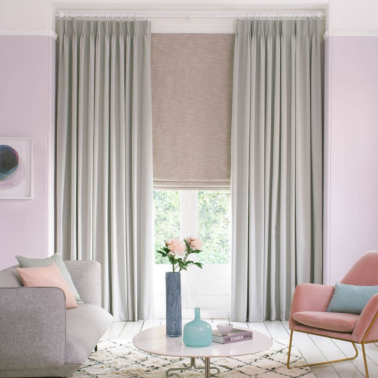 Floor-length pleated grey curtains frame a lowered beige Roman blind; drawn to the sides while the blind filters light, revealing a leafy view in a softly lit pastel living room.

THE FASHION BOOK
