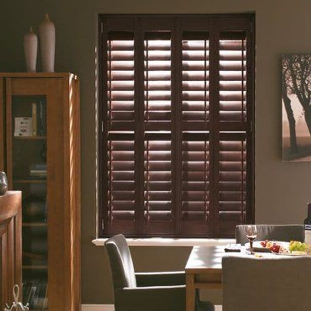 Dark wooden plantation shutters covering a central window, slatted louvers partially angled to filter sunlight; in a warm-toned dining room with wooden furniture, a set table, cabinet and wall art.