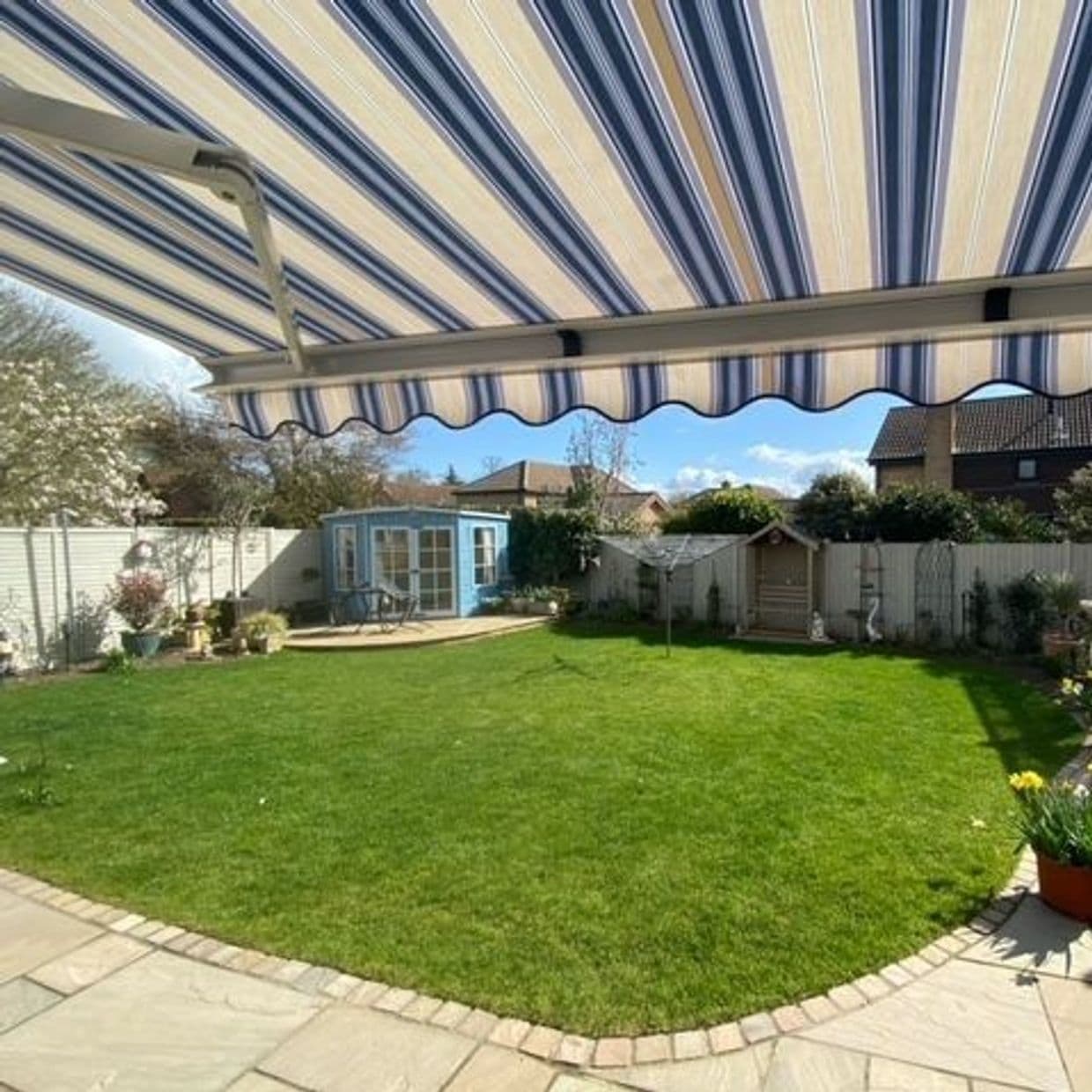 Striped blue-and-cream retractable awning, fully extended with scalloped valance, shading a patio overlooking a sunny fenced backyard with green lawn, potted plants, and a small blue summerhouse.