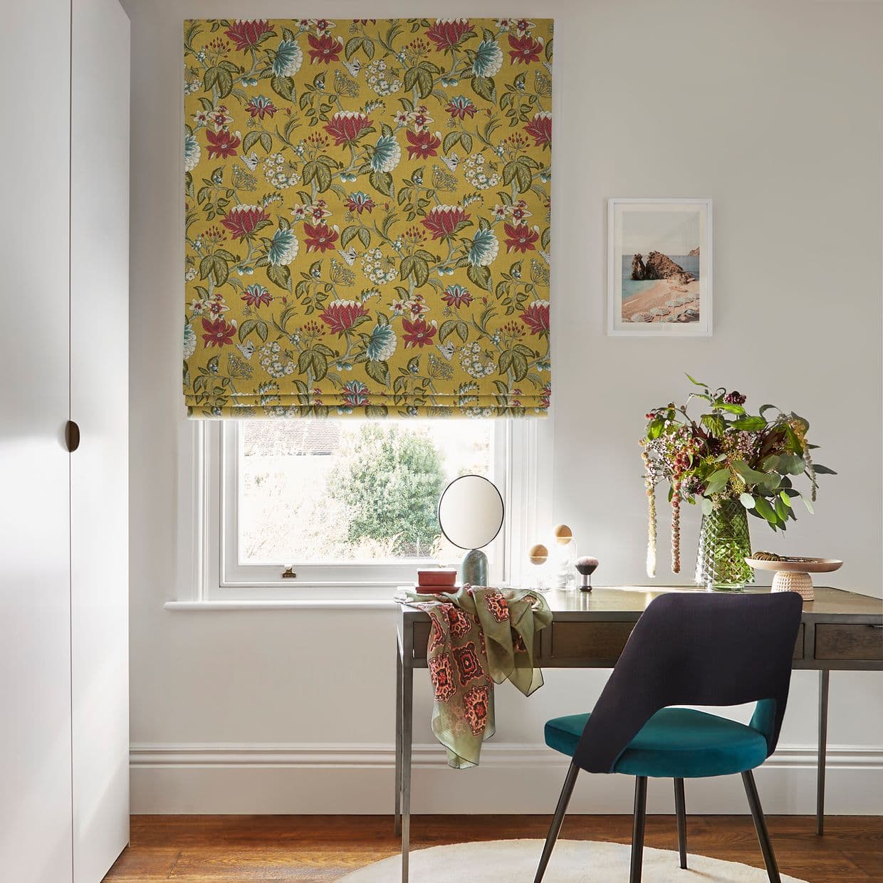 Roman blind with mustard-yellow floral print, lowered with soft folds, filtering daylight through window; tidy desk and teal chair beneath, vase of greenery, pale walls and framed seaside print.