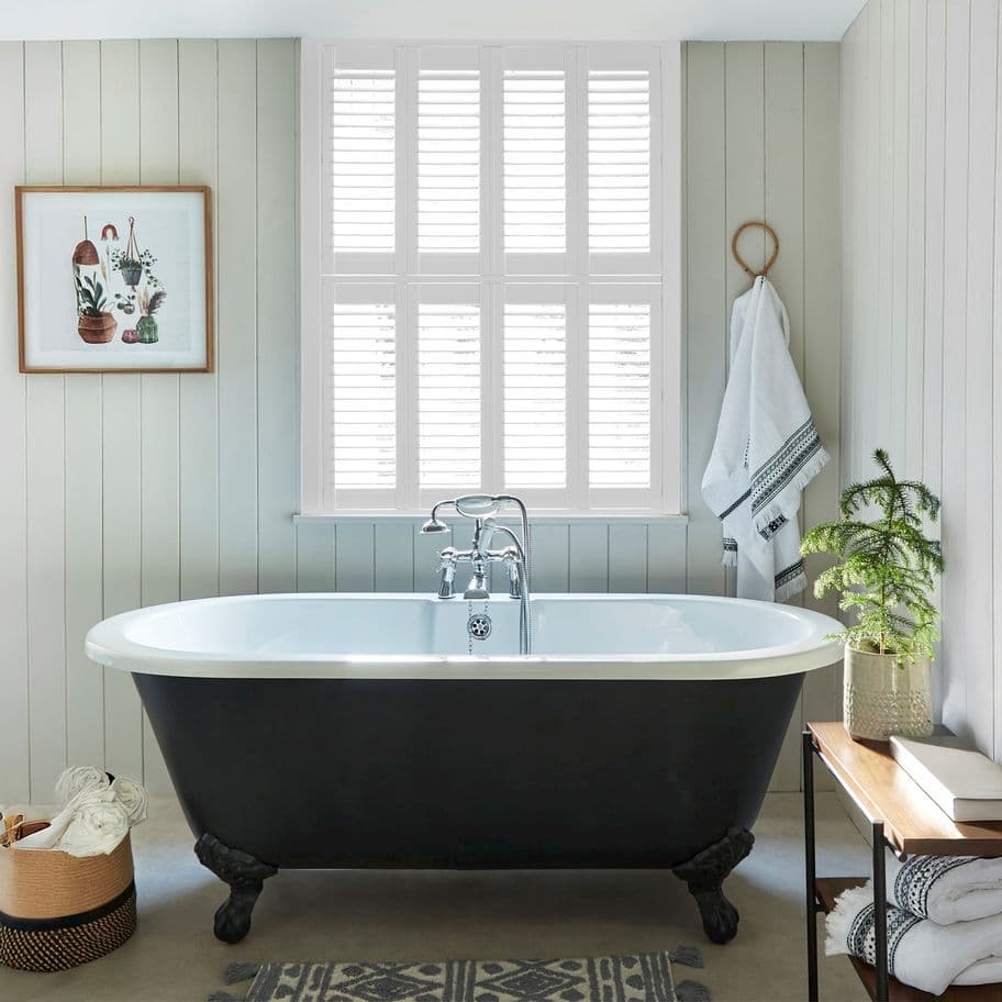 White plantation shutters covering a tall window, slats closed and filtering bright daylight; set above a black clawfoot bathtub in a lightâ€‘paneled, softly lit bathroom with towel and potted plant.