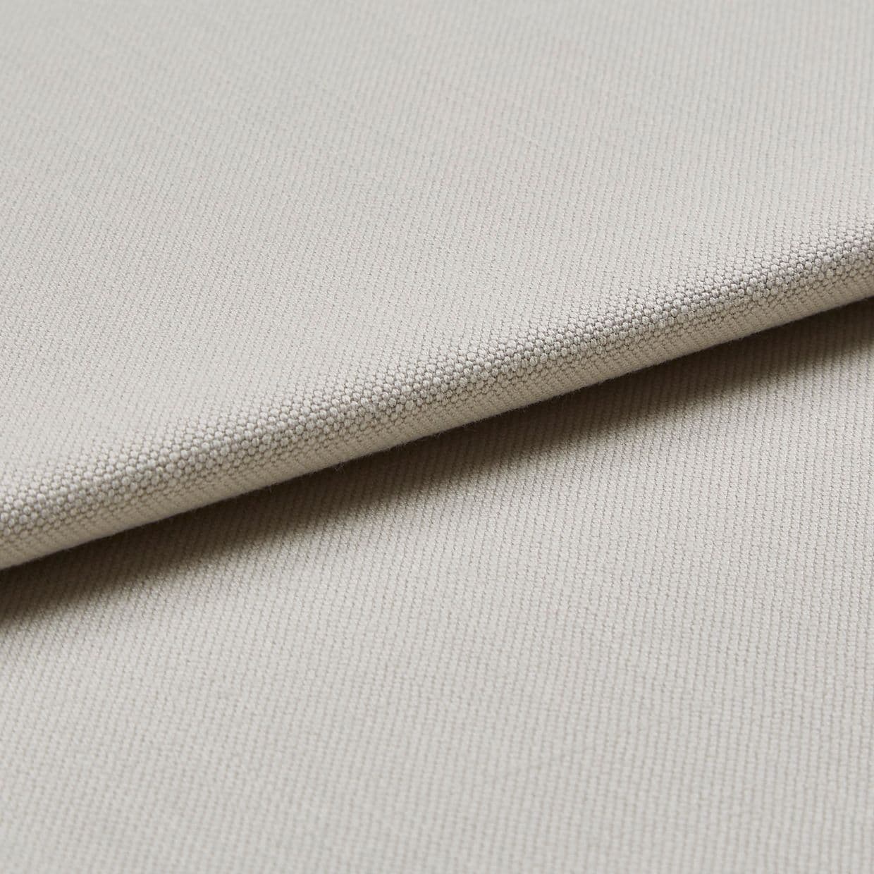 Beige woven fabric swatch folded diagonally, showing tight pebble-like texture; layered close-up under soft, neutral lighting, suggesting upholstery or curtain material.