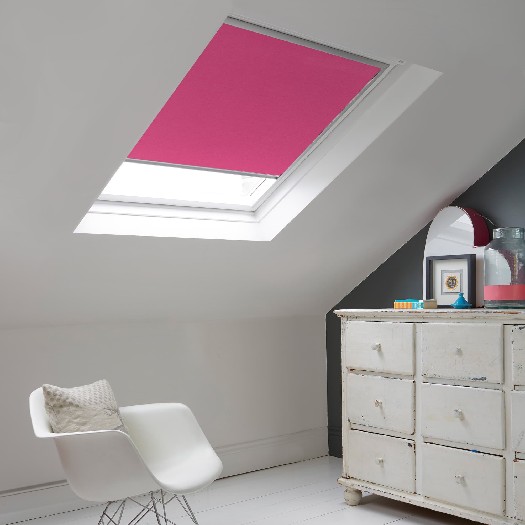Room with white eaves wall, dark blue-grey wall to right with a shabby chic white drawer unit, a contemporary white chair, white painted floorboards and a skylight blind in hot pink.